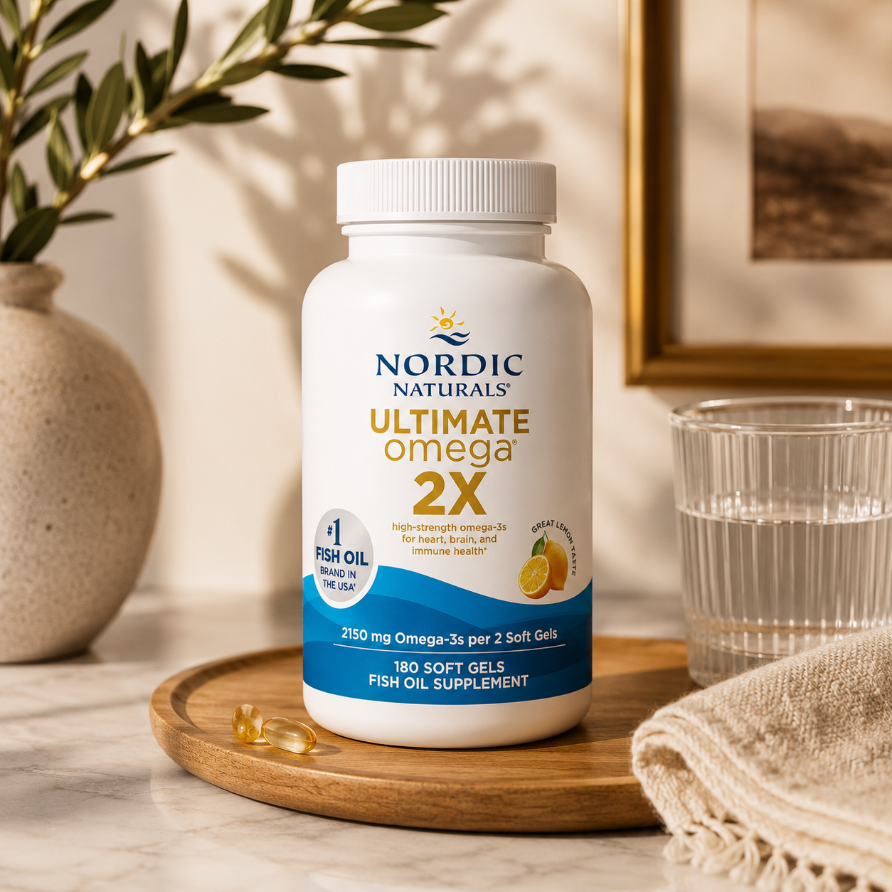Ultimate Omega 2X fish oil supplement bottle displayed on a wooden tray with a glass of water, a beige cloth, and olive branches in a warm, styled setting.