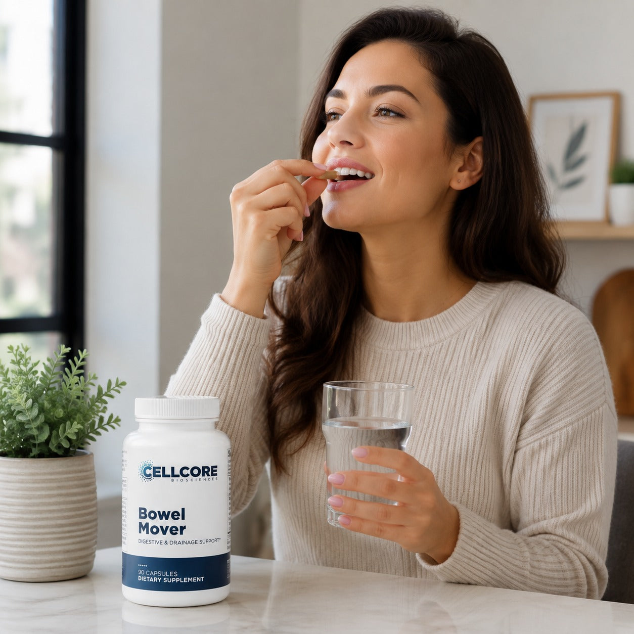 A woman in a cream ribbed sweater is shown taking a pill from her finger while holding a glass of water. In the foreground, on a white table, is a white bottle of  Bowel Mover dietary supplement capsules.